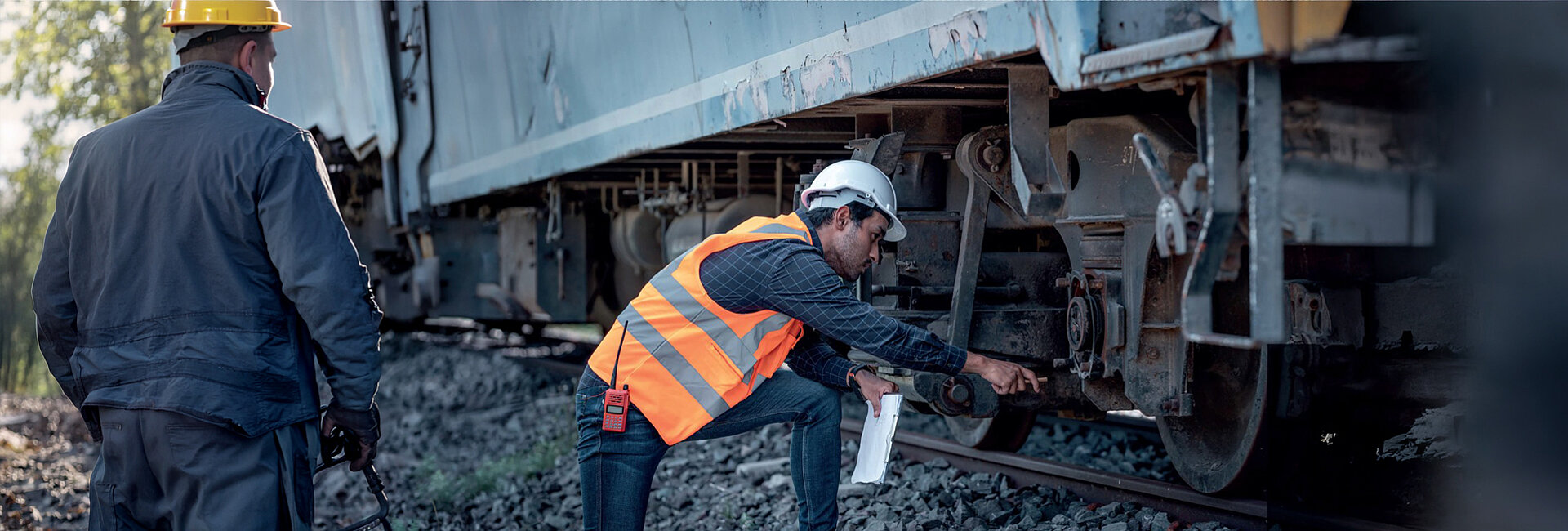 Zwei Bahnmitarbeiter in Schutzausrüstung inspizieren auf einem Bahnsteig eine entgleiste oder beschädigt aussehende Bahreuse. Einer bückt sich, um die Räder und Schienenverbindung zu prüfen, beide tragen Warnwesten bzw. Helm und arbeiten im Gleisbett.