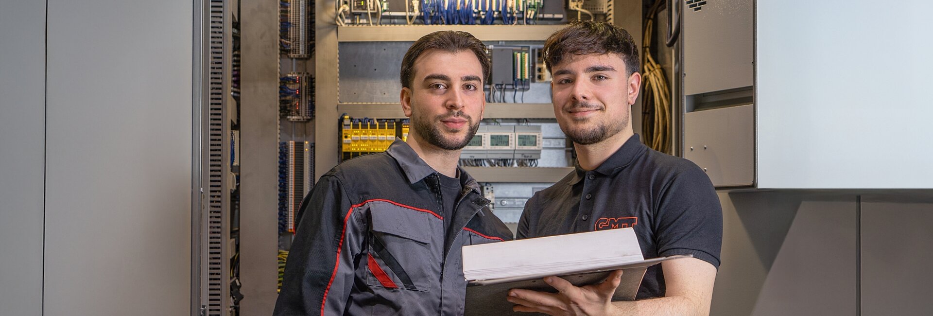 Zwei Nachwuchskräfte arbeiten vor einem geöffneten Schaltschrank im technischen Bereich.