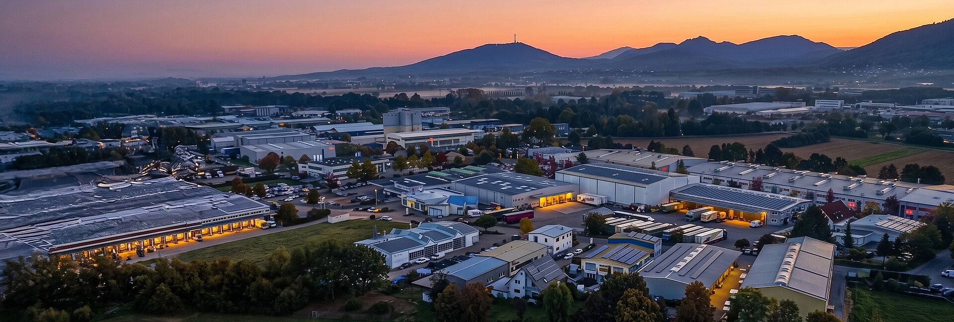 Groß Britanische Industrieanlage bei Sonnenuntergang mit weitläufigen Hallen, Lagerbereichen und Verwaltungsgebäuden, umgeben von sanften Hügeln und in der Ferne liegendem Gebirge.

(49 Wörtern)