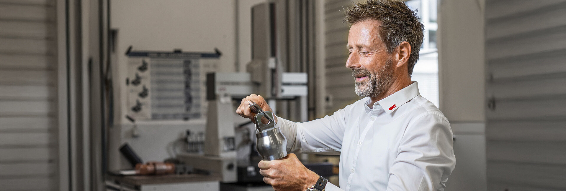Ein Mann in weißem Oberteil und grauer Schürze prüft konzentriert eine Flasche in einem Labor oder Produktionsbereich mit Technikgeräten und Regalen im Hintergrund.