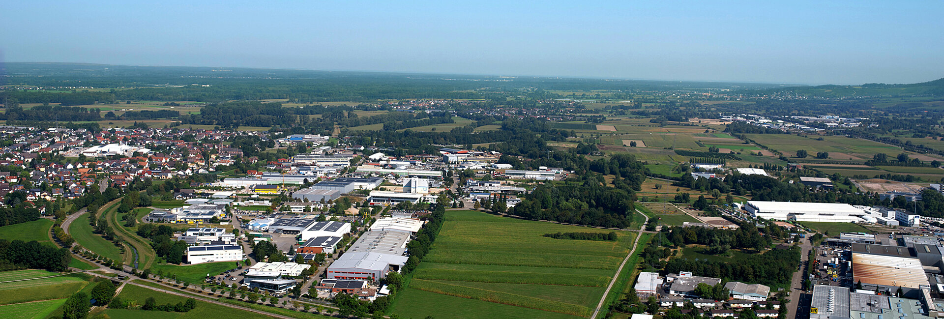 Aerial view showing an industrial area surrounded by green fields, small villages with clusters of buildings, and roads winding through the suburban landscape with patches of forest on the right.